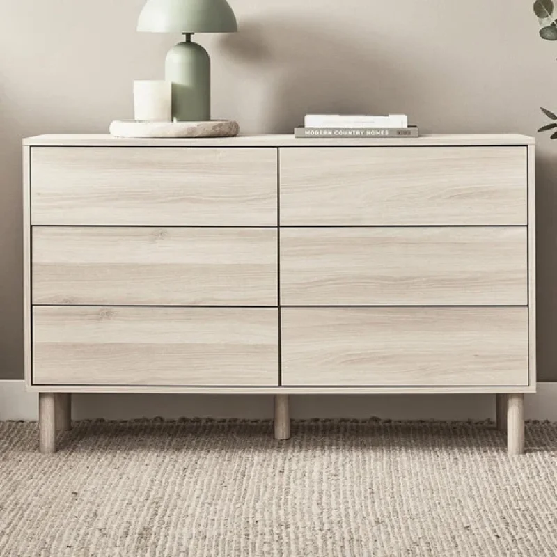 A light wood 6-drawer chest with tapered legs, styled with a lamp and books on top.