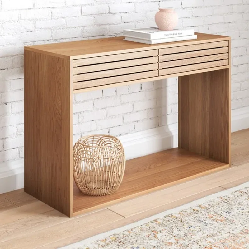 A light oak slatted console table with two drawers and an open shelf below, styled with books and a vase on top, and a wicker basket on the shelf.