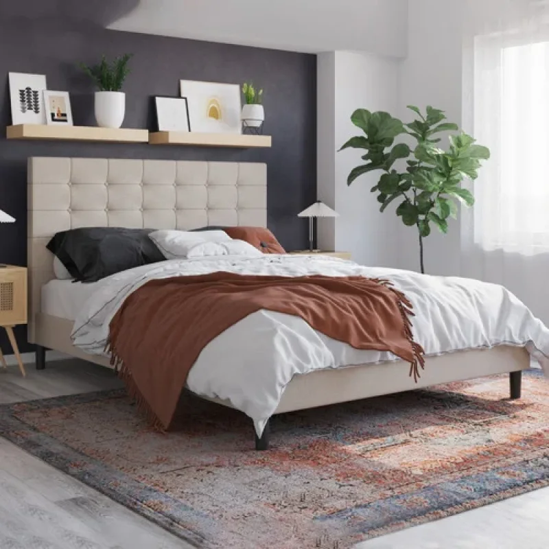 A beige upholstered bed frame with a button-tufted headboard, dressed with white and black bedding and a rust-coloured throw, in a stylish bedroom setting.