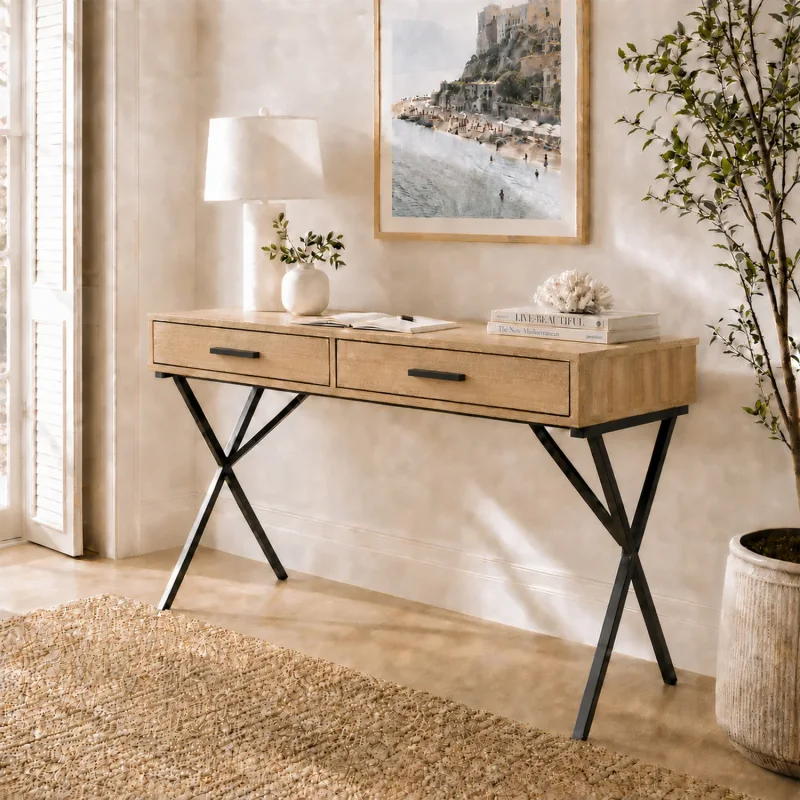 A light wood grain desk with two drawers and black X-shaped legs, styled in a room with a lamp, books, and a plant.