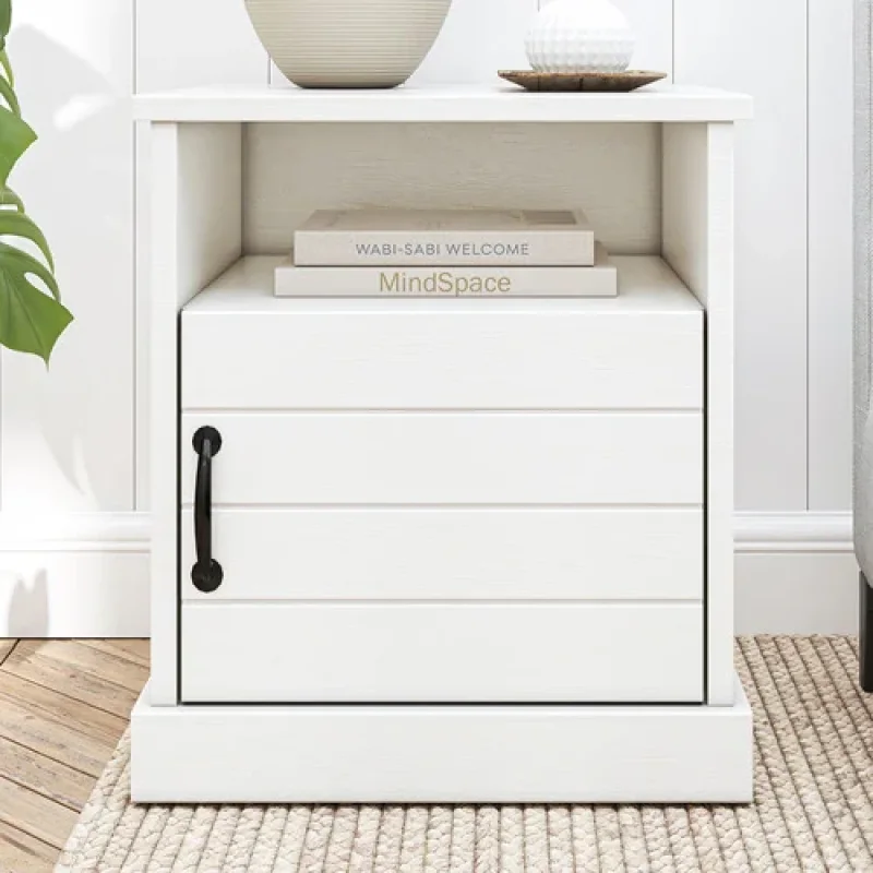 A white Hamptons-style bedside table with a slatted door, a drawer, and an open shelf, featuring a black handle and a textured rug underneath.