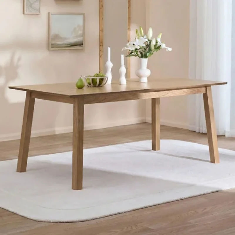 A 180cm natural oak wood dining table with a light wood grain veneer top and angled legs, set on a white rug in a minimalist dining room.