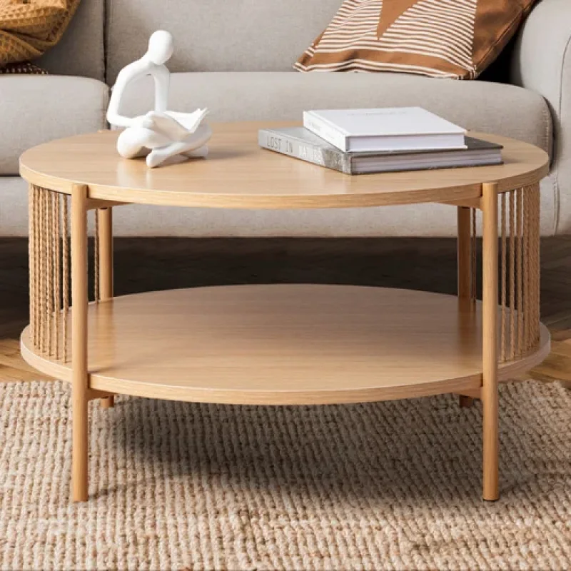 A round, light wood coffee table with two tiers and cane detailing on the sides, placed on a woven rug in a living room setting.