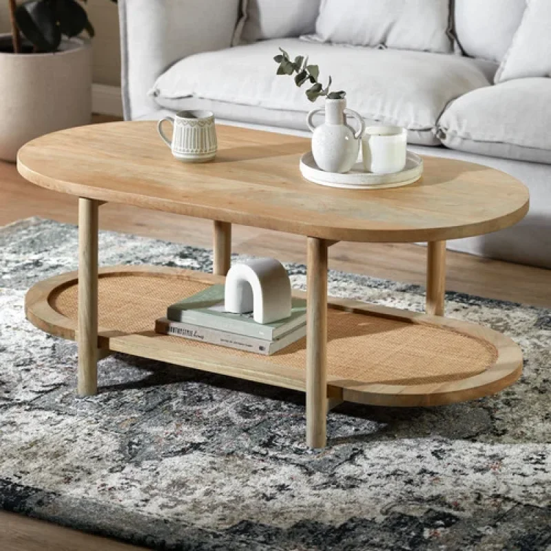 An oval-shaped coffee table made of natural mango wood with a lower shelf featuring cane webbing, placed on a patterned rug in a living room setting.