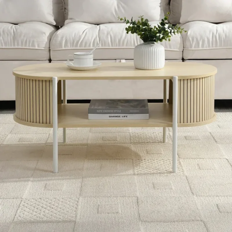 A light wood oval coffee table with a slatted design on the sides and a lower shelf, placed on a cream rug in front of a white sofa.