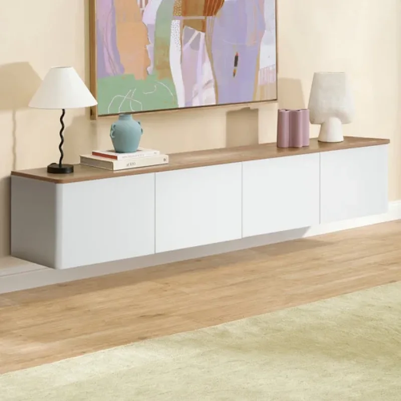 A long, white, wall-mounted TV unit with a timber-look top, featuring four doors and mounted on a wall above a light-coloured rug.