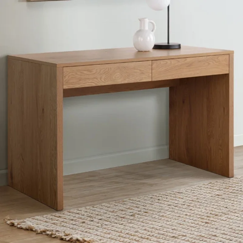 A light oak veneer writing desk with two drawers and a minimalist design, shown in a room setting with a lamp and a small jug on top.