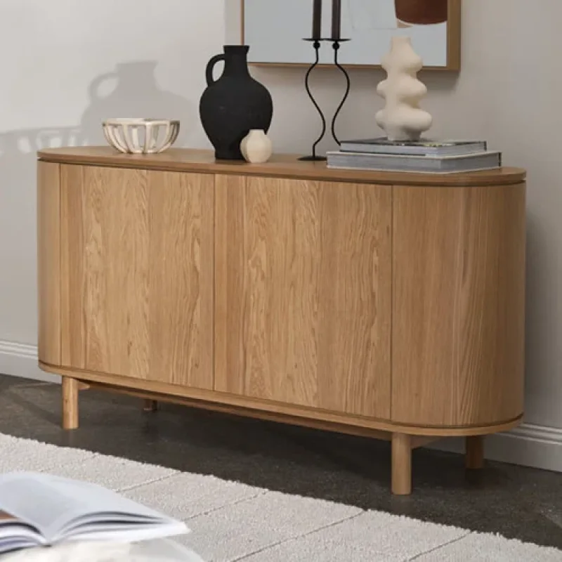 A light oak timber sideboard with a curved front and four doors, standing on slim tapered legs. It is styled with decorative items on top.
