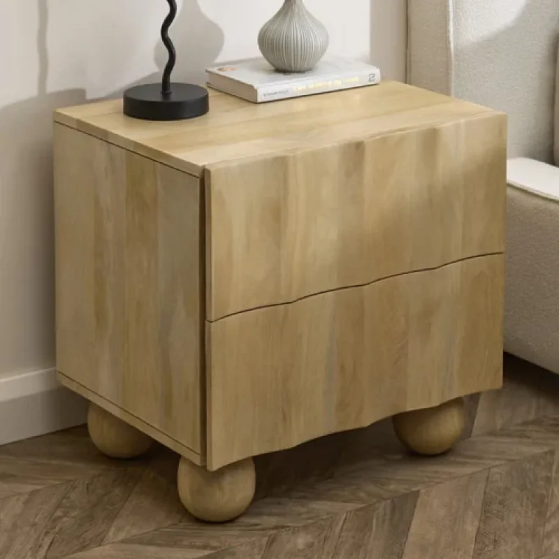 A light-coloured mango wood bedside table with two drawers and rounded legs, featuring a wavy design on the drawer fronts. A lamp and a book are placed on top.