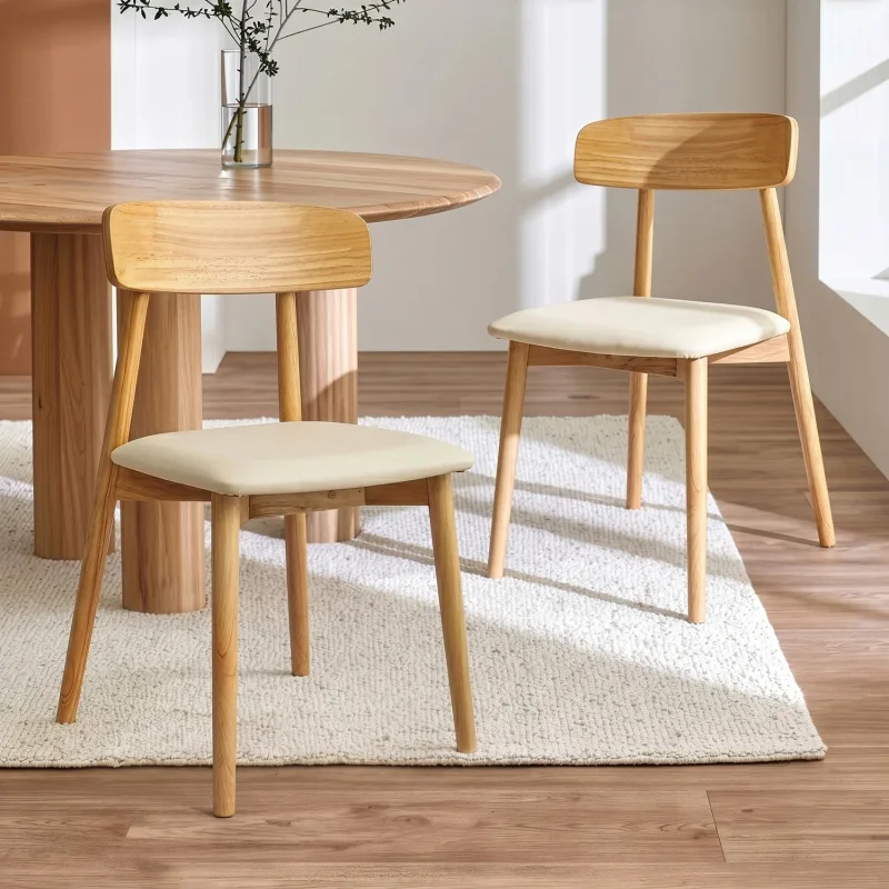 Two Sujin Rubberwood Dining Chairs with cream upholstered seats and natural wood frames, placed on a textured rug next to a round wooden dining table.