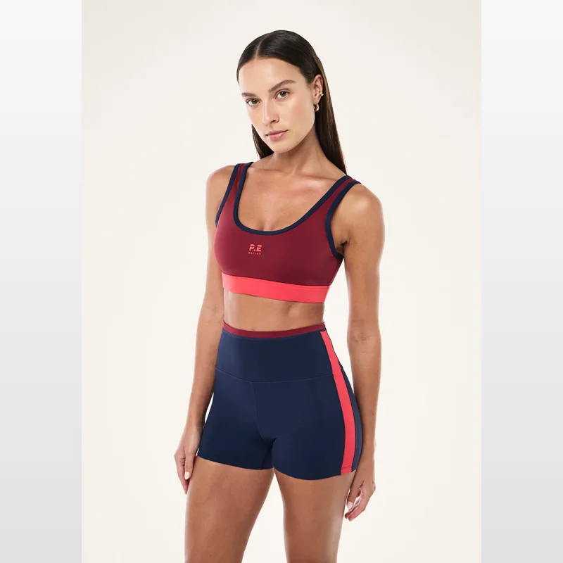 Woman wearing a burgundy Pe-nation Pursuit Sports Bra with navy trim and a coral band, paired with matching navy shorts with a coral stripe.