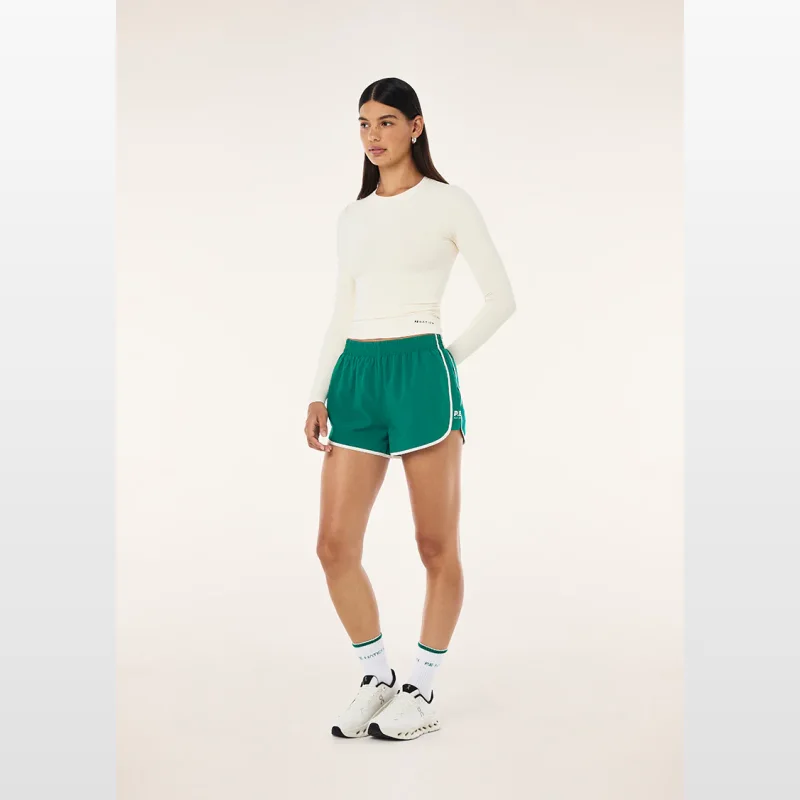 Woman wearing Pe-nation Preset Shorts in College Green with Whisper White trim, paired with a cream long-sleeve top and white sneakers.