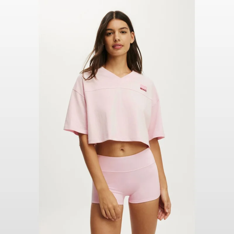 Woman wearing a pink cropped graphic sporty tee with a v-neck and dropped shoulders, paired with pink shorts.