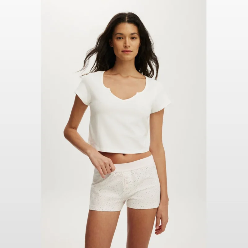 Woman wearing a white cropped short-sleeve sleep tee with a notched neckline and light pink trim on the waistband of her polka dot shorts.