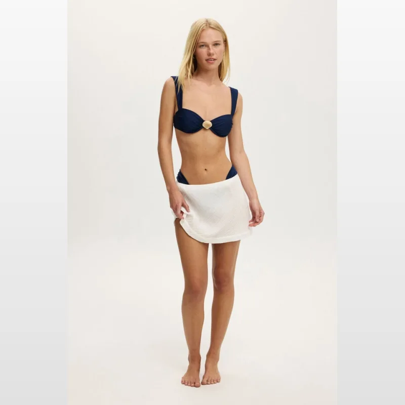 A blonde woman wearing a navy blue bikini top and a white textured mini skirt, posing against a white background.