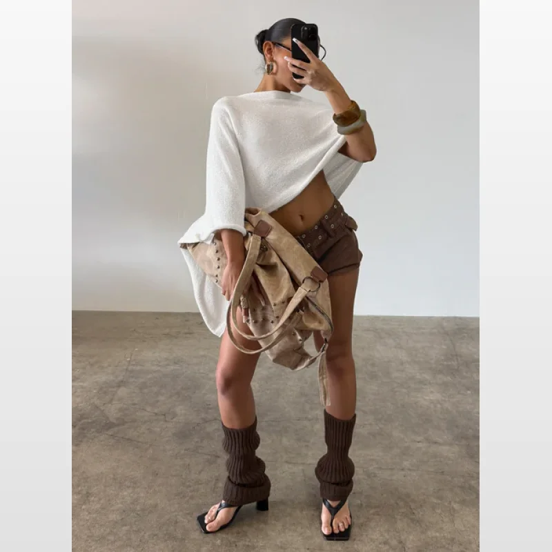 Woman wearing a white off-shoulder knit poncho, brown shorts, brown leg warmers, and tan studded shoulder bag.