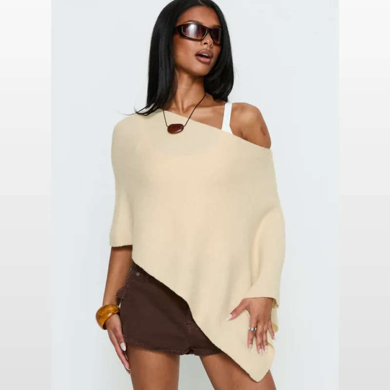 Woman wearing a cream off-shoulder heavy knit poncho with a white tank top underneath and brown shorts.