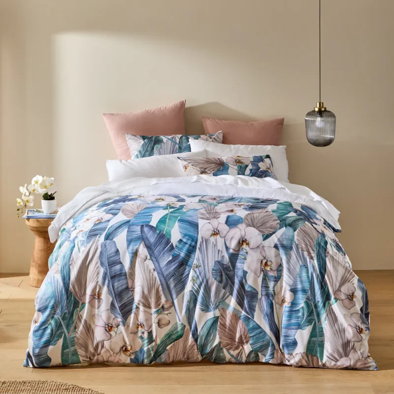 A bed dressed in the Adorn Living Rowie Quilt Cover Set, featuring a vibrant tropical print of palm leaves and orchids in blues, teals, and blush tones.