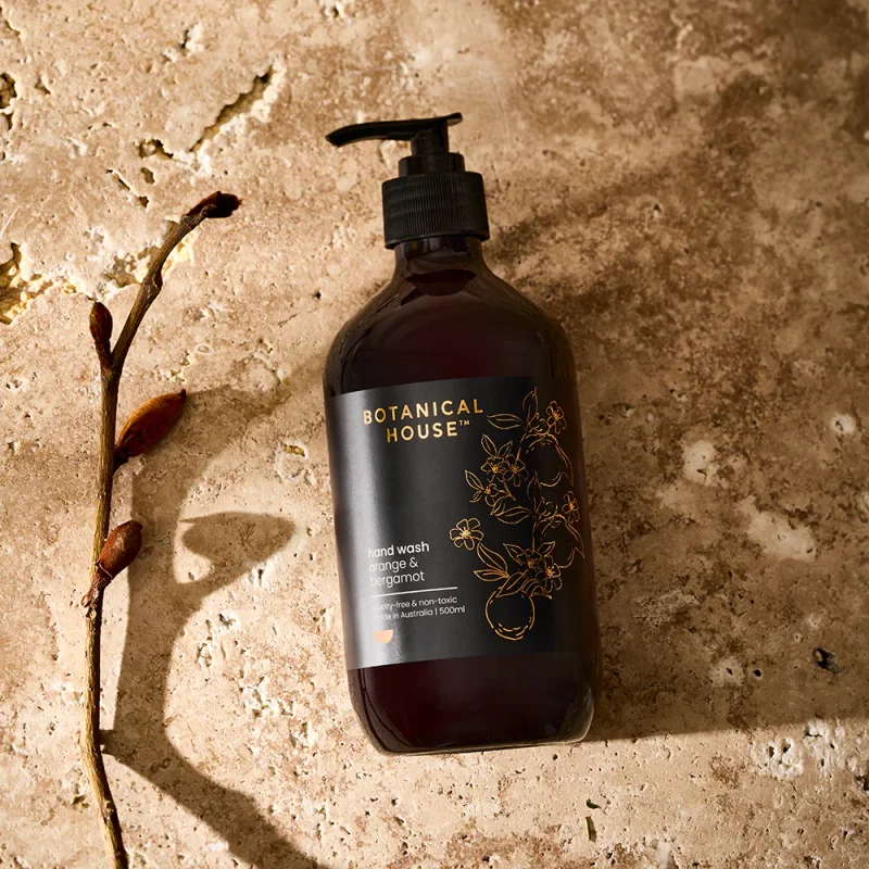 A dark amber bottle of Botanical House Orange & Bergamot Hand Wash with a black pump dispenser, set against a textured beige background with a sprig of dried foliage.