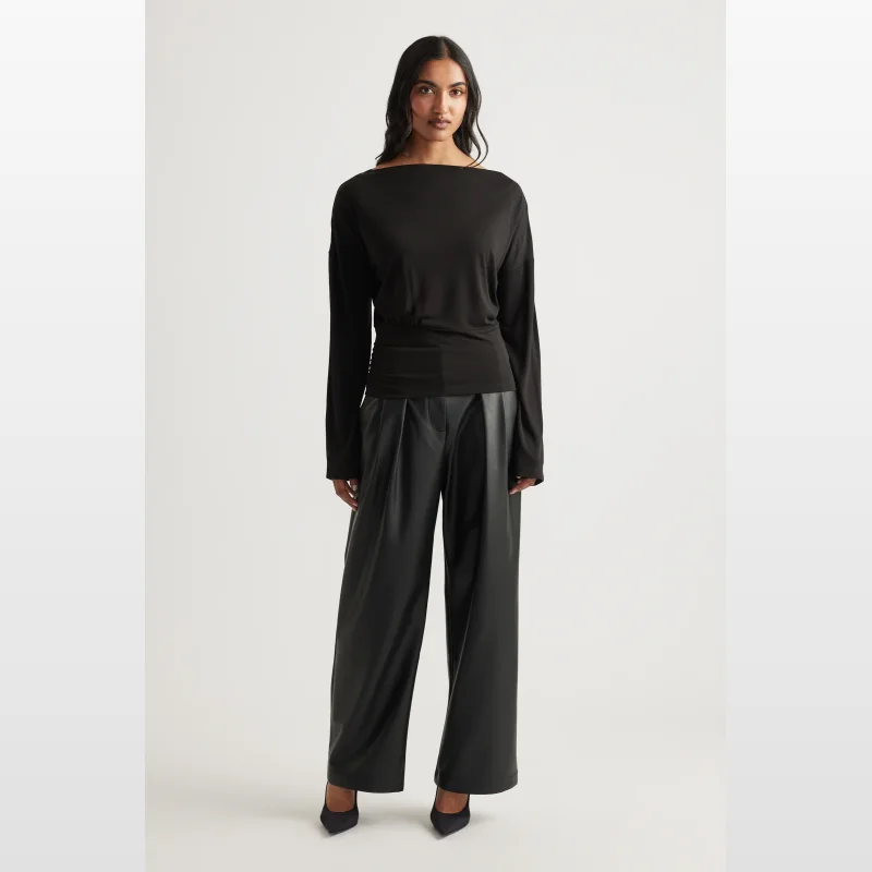 Woman wearing a black long sleeve top with a boat neck and cinched waist, paired with wide-leg black pants.