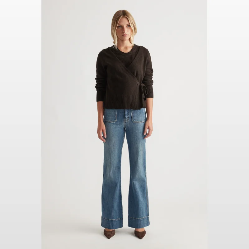 Woman wearing a dark brown long-sleeved wrap knit jumper with a built-in tank top and tie detail at the side, paired with wide-leg blue jeans.