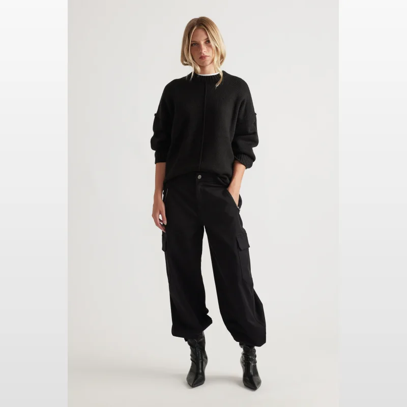 Woman wearing a black long-sleeved knit jumper with seam detailing and ribbed trims, paired with black cargo pants and black heeled boots.