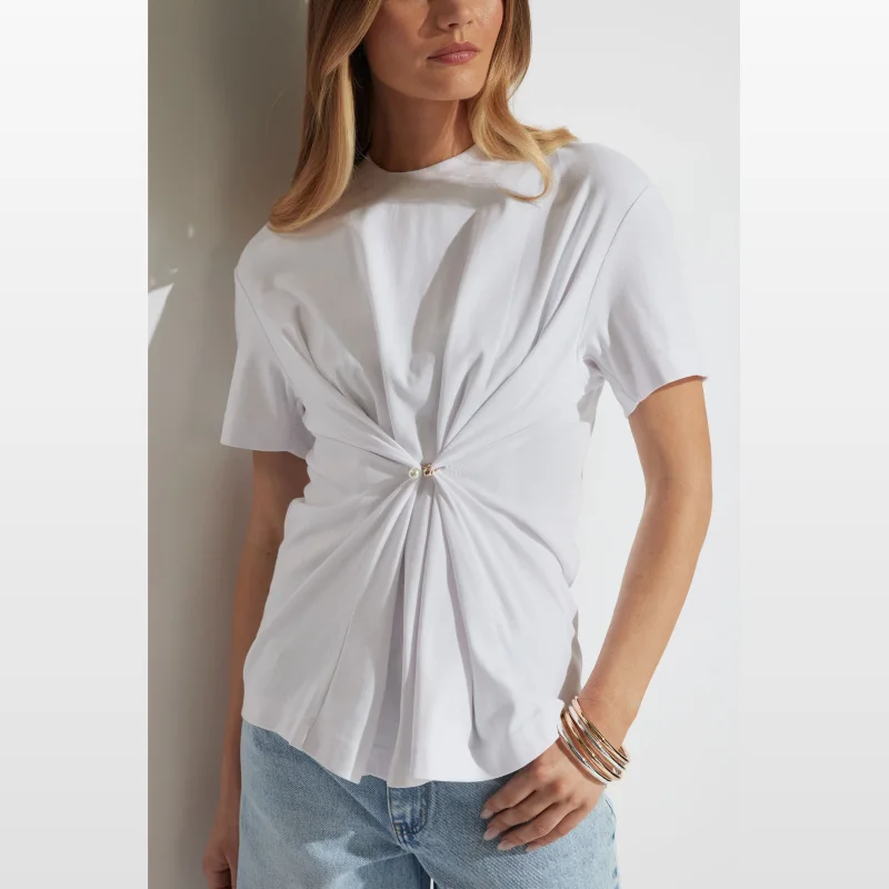 Woman wearing a white oversized t-shirt with a twisted front detail secured by a pearl and gold-tone metal clasp, crew neckline, and short sleeves.