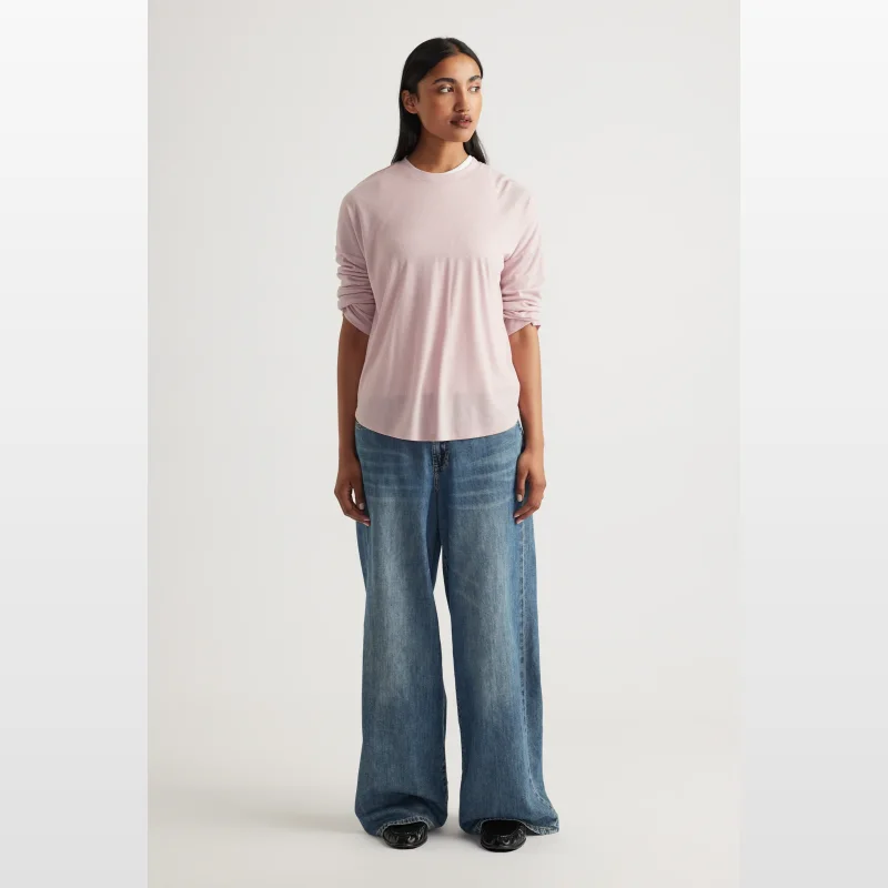 Woman wearing a light pink long-sleeve ribbed crew neck top with a scoop hem and drop shoulders, paired with wide-leg blue jeans.