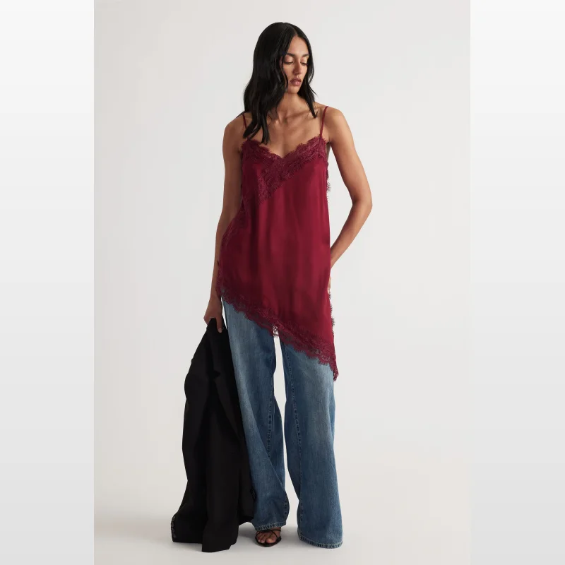 Woman wearing a burgundy lace-trimmed tunic with thin adjustable straps and an asymmetric hem, paired with blue jeans and holding a black blazer.