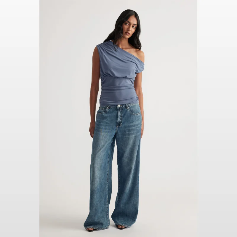 Woman wearing a blue Decjuba Sienna off-shoulder top with an asymmetrical neckline and gathered side seams, paired with wide-leg jeans.
