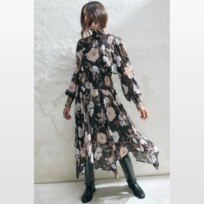 Woman wearing a black floral print midi dress with full length puff sleeves, a raised shirred neckline, shirred waist, and handkerchief hem.