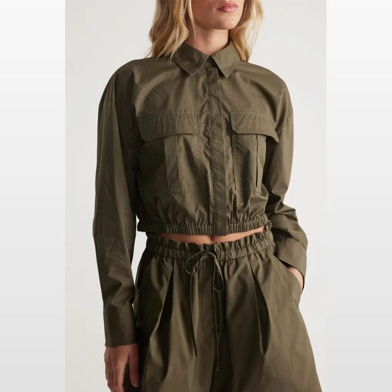 Woman wearing a khaki green Decjuba Rey Crop Shirt with long sleeves, collared neckline, patch pockets, and an elasticated cropped hem.