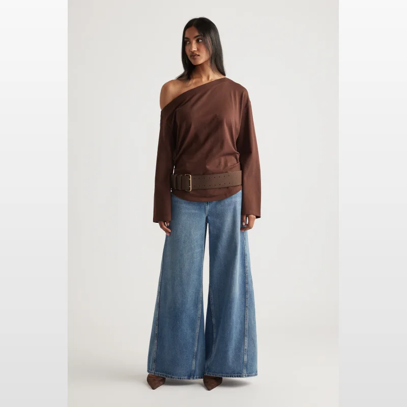 Woman wearing a brown long-sleeved Ally Tipped Shoulder Tee with an asymmetrical neckline and gathering at the side waist, paired with wide-leg denim jeans and a brown belt.