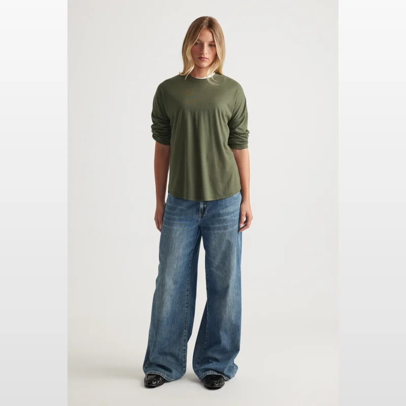 Woman wearing a green long sleeve ribbed crew neck top with a scoop hem and drop shoulder, paired with wide-leg blue jeans.