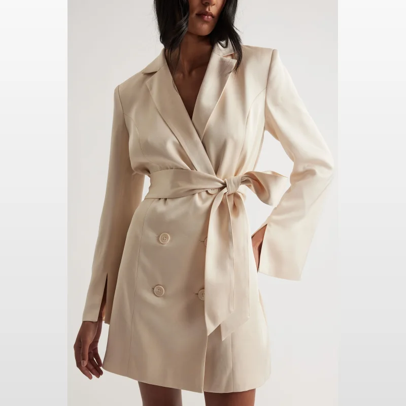 Woman wearing a cream satin Celiene blazer dress by Decjuba, featuring a collared neckline, double-breasted front, detachable waist tie, and midi length.
