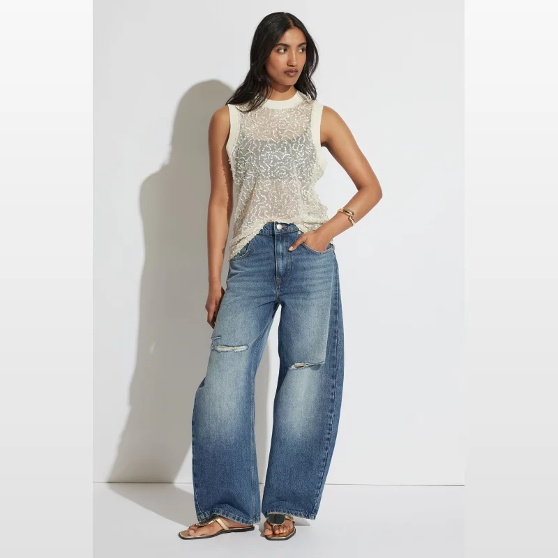 Woman wearing a cream-coloured, sleeveless sequin tank top with a crew neckline and ribbed trim, paired with distressed blue denim jeans.