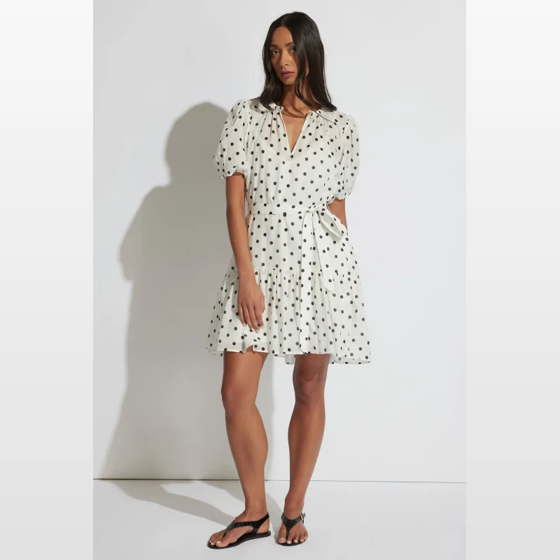 Woman wearing a white Dalia Mini Dress with black polka dots, featuring puff sleeves and a tie waist.
