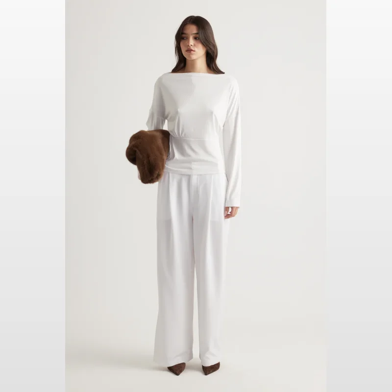 Model wearing a white long-sleeved boat neck top with a cinched waist and drop shoulders, paired with white wide-leg pants.