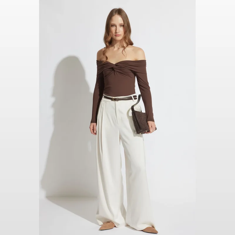 Woman wearing a brown long sleeve off-shoulder top with a twist front detail, paired with white wide-leg trousers and a brown belt.