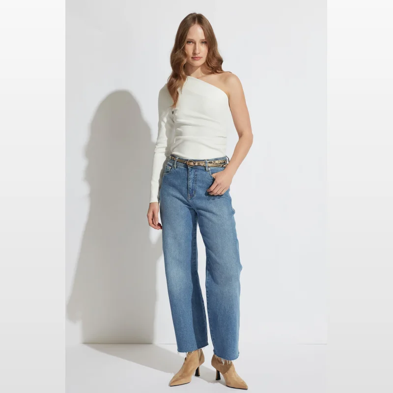 Woman wearing a white one-shoulder long-sleeved top with ruched detailing at the side seam, paired with blue denim jeans and tan heeled boots.