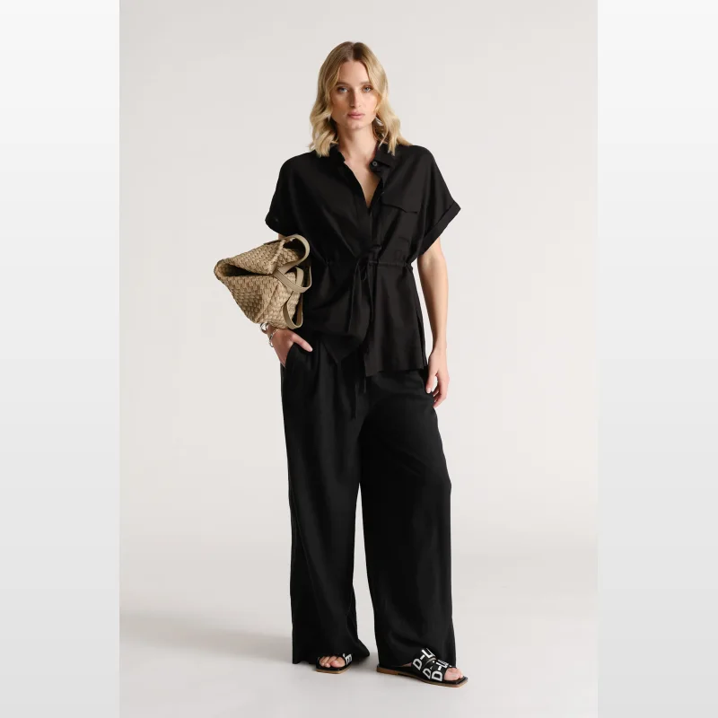 Woman wearing a black D-Luxe Linen Blend Short Sleeve Shirt with a tie waist and patch pockets, paired with black wide-leg pants and black slides.