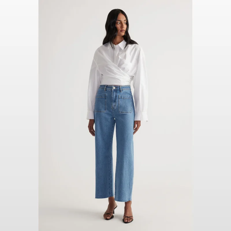 Model wearing Decjuba Darcy high rise wide leg ankle length jeans in blue stretch denim, paired with a white wrap shirt.