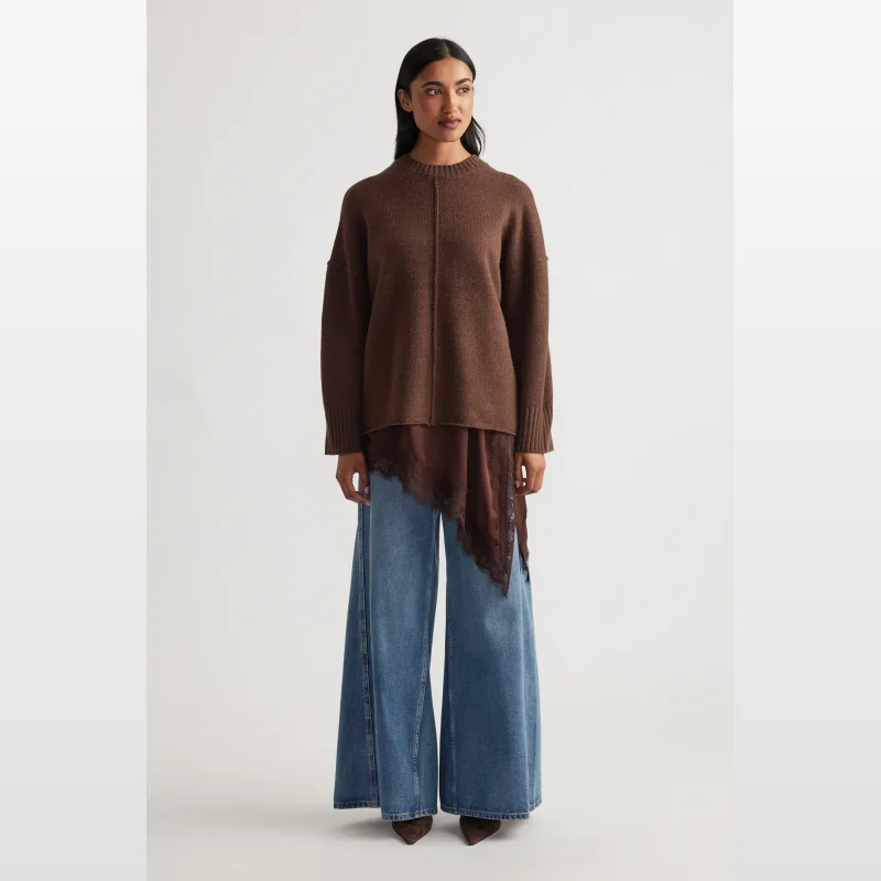 Woman wearing a brown long-sleeved knit jumper with seam detailing and ribbed trims, paired with wide-leg blue jeans and brown pointed-toe heels.