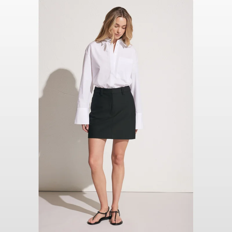 Model wearing a black Decjuba Alex mini skirt with a white button-up shirt and black sandals.