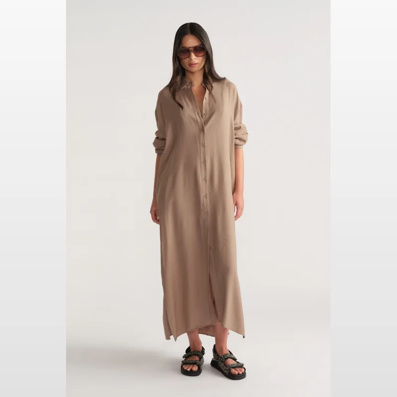 Woman wearing a long, collared, button-through shirt dress in a textured tan fabric, with long sleeves and side splits.