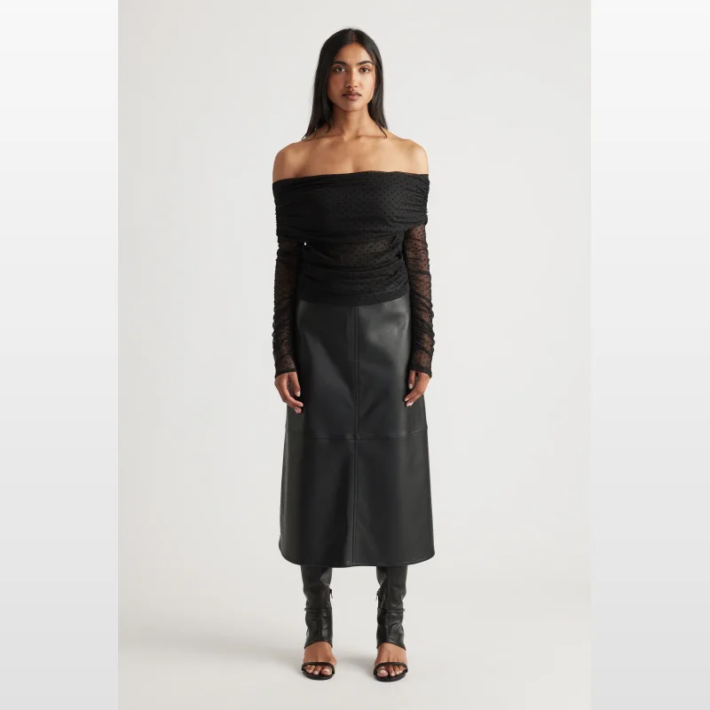 Woman wearing a black off-shoulder ruched mesh top with polka dot pattern and long sleeves, paired with a black leather midi skirt and black heeled sandals.