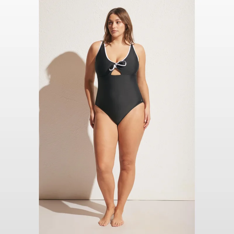 Woman wearing a black Decjuba one-piece swimsuit with a contrast white trim and a tie front detail.
