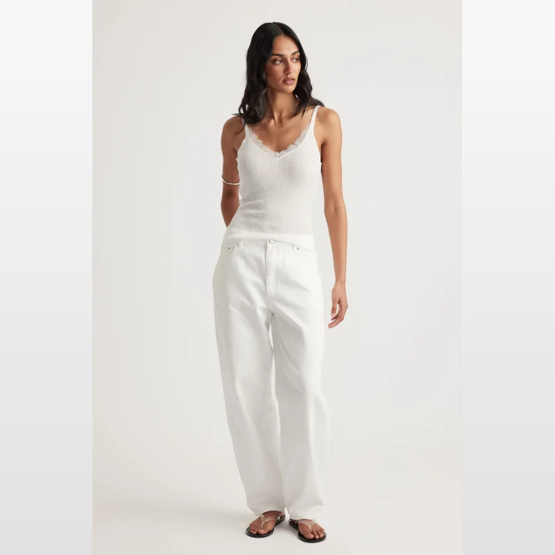 Woman wearing a white ribbed knit tank top with a V-neckline trimmed in lace, and adjustable straps. She is also wearing white wide-leg jeans.