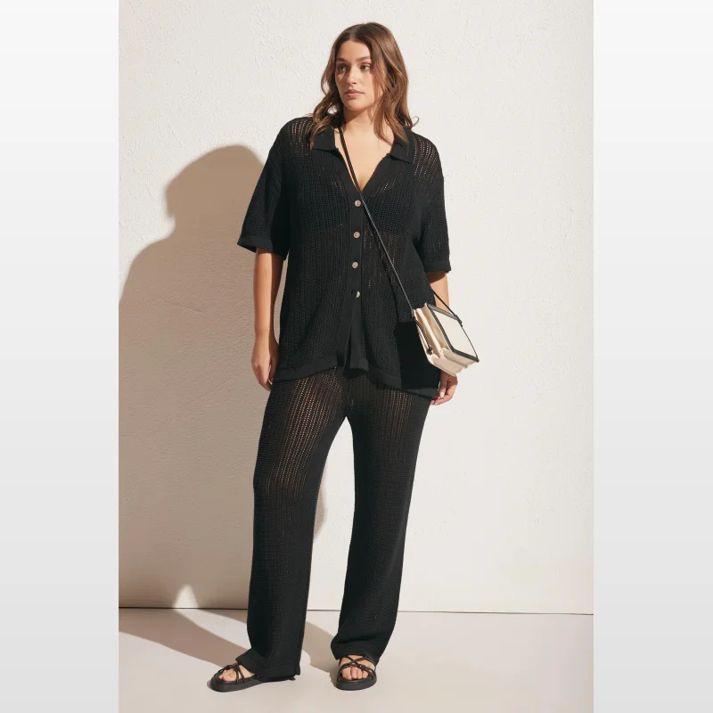 Woman wearing black crochet wide-leg pants and a matching short-sleeved collared shirt, with a cream and black crossbody bag.