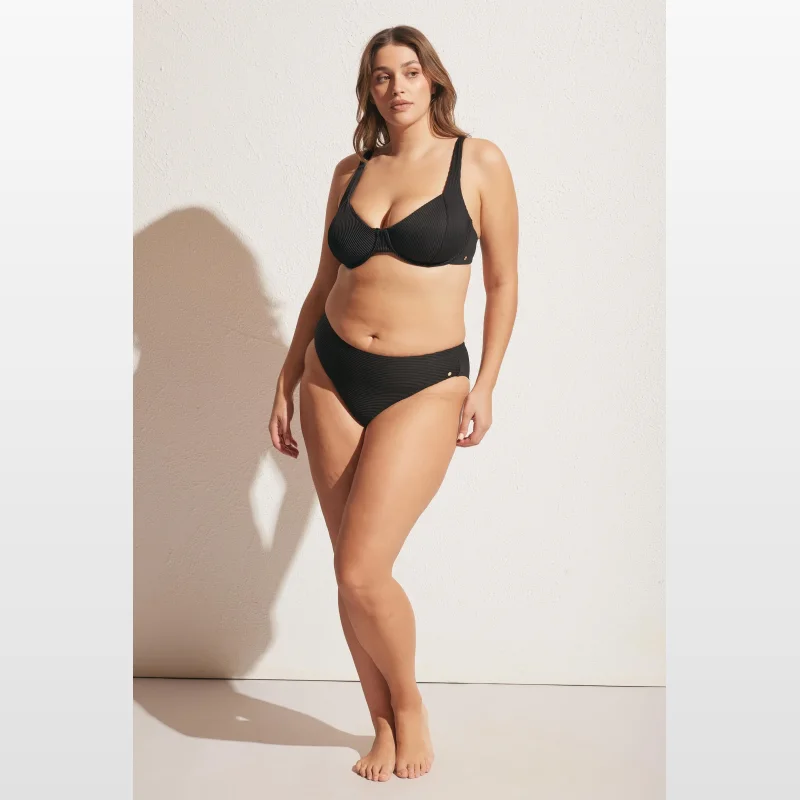 Woman wearing black ribbed bikini bottoms with medium brief coverage and medium rise, featuring gold metal hardware detailing.